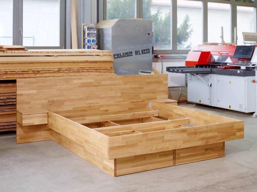 Massivholzbett mit Stauraum in einer Werkstatt mit verschiedenen Holzbrettern und Maschinen